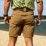 Rear view of a man wearing tan Field Shorts standing on a sandy shore with water and greenery in the background, showcasing the casual and durable design of the shorts.