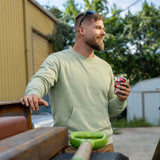 Man wearing a light green Smuggler French Terry Crew sweatshirt, smiling outdoors while holding a can, leaning on a wooden surface in a casual setting.