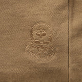 Close-up of a brown fabric with an embroidered skull and spade emblem and the text "SMUGGLER," part of the design on the Smuggler French Terry Crew sweatshirt.