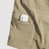 Close-up of a tan pair of Field Shorts showing a zippered side pocket partially unzipped with a white electronic device inside.