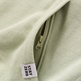 Close-up of a light sage green Smuggler French Terry Crew showing a hidden zipper pocket and a fabric tag with the text "STAY ZERO" on textured fabric.