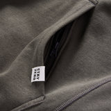 Close-up of an olive green Smuggler French Terry Crew showing a zippered pocket detail and a fabric tag with the words "STAY ZERO.