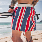 Man wearing Freedom Stripe Swim Trunks with red, white, and blue vertical stripes, standing on a sandy beach holding a surfboard near the ocean.