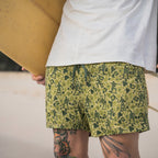Man wearing green patterned Swim Trunks with small dark silhouettes of people, standing outdoors holding a surfboard, with visible leg tattoos.