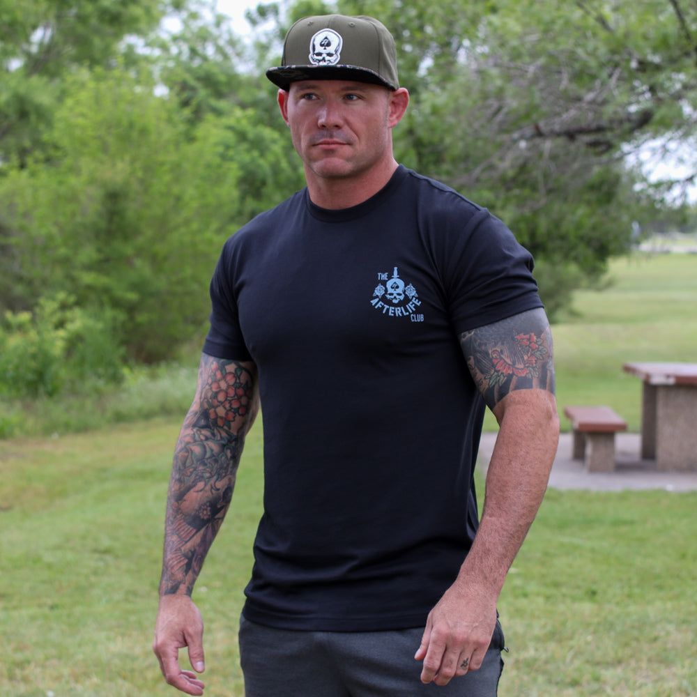 Clever Girl Tee black short sleeve shirt with a small skull graphic worn by a tattooed man outdoors in a grassy park setting