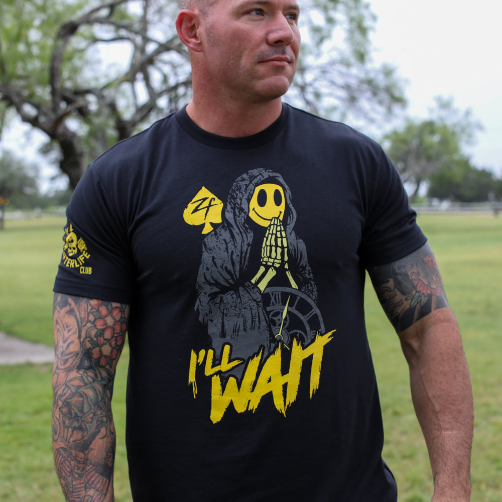 I'll Wait Tee black graphic T shirt featuring a yellow hooded skeleton design and bold yellow lettering, modeled outdoors in a park