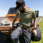 Man wearing an olive green ZF Core Tee with tactical gear, sitting on the back of an off-road vehicle in a grassy outdoor setting under a clear sky.