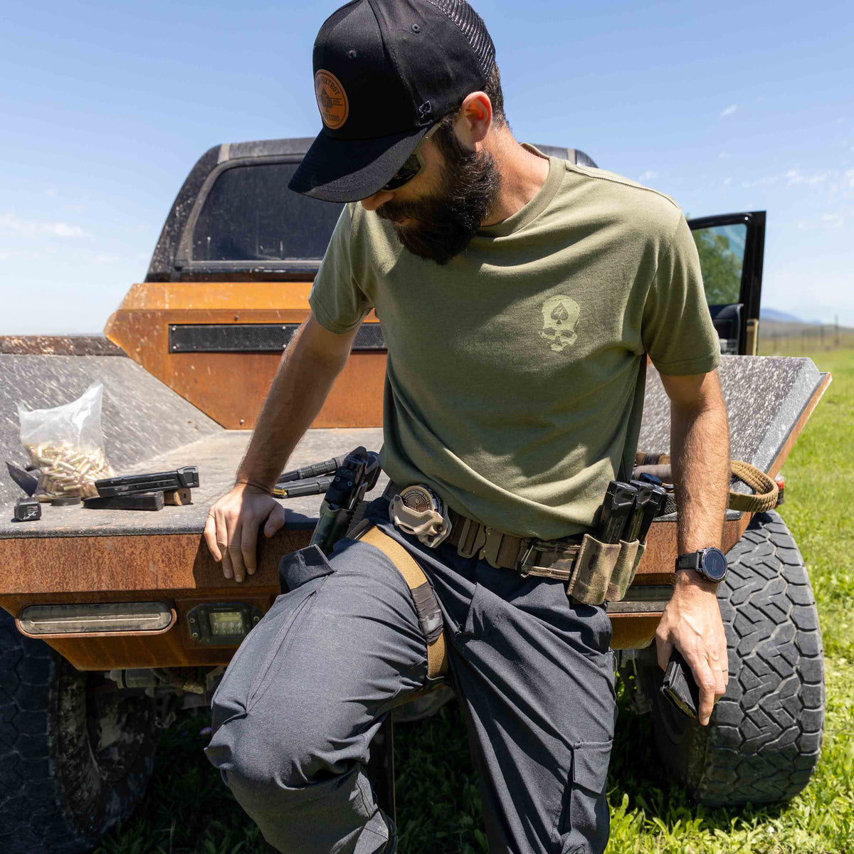 Man wearing an olive green ZF Core Tee with tactical gear, sitting on the back of an off-road vehicle in a grassy outdoor setting under a clear sky.