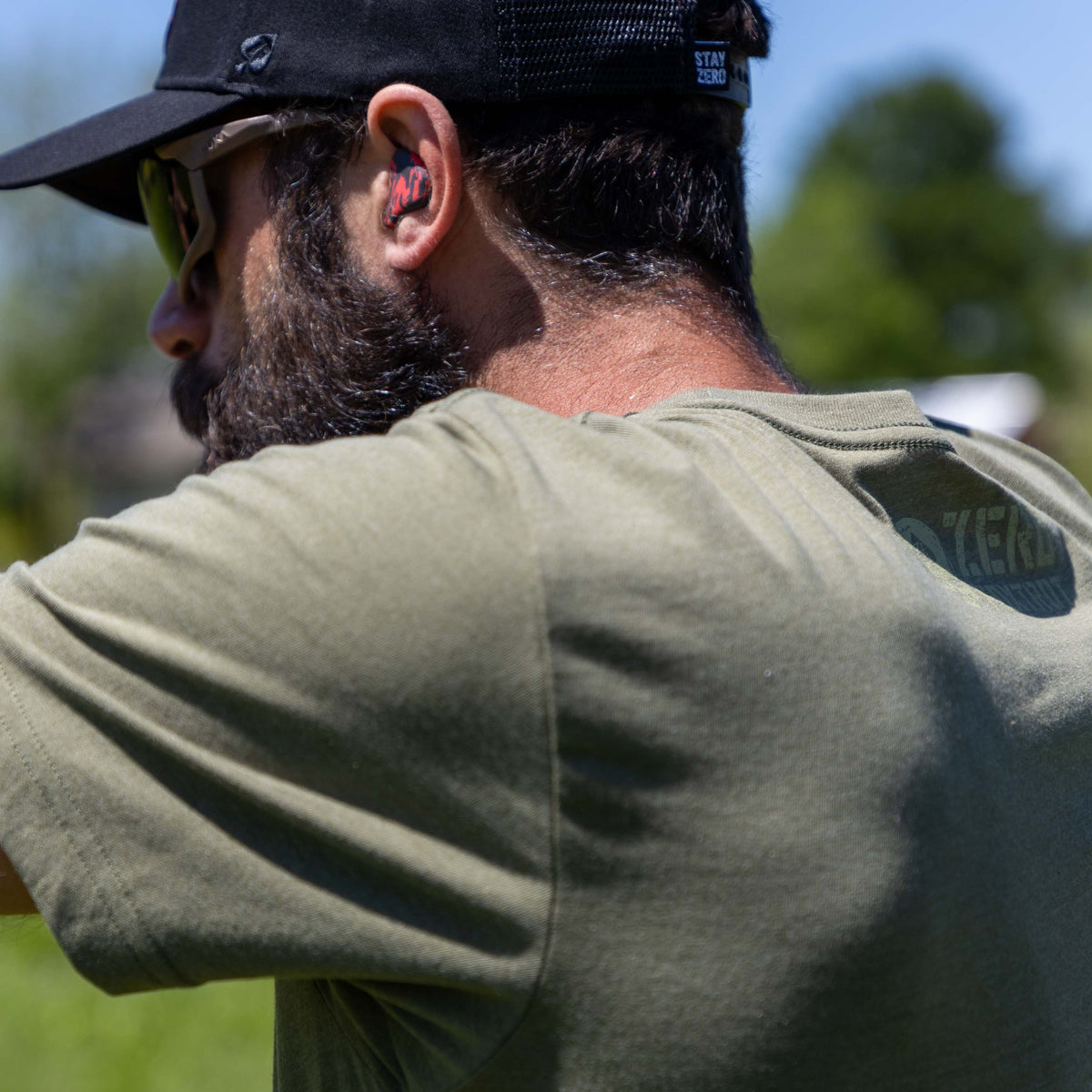 Man wearing an olive green ZF Core Tee shirt outdoors, shown from the back with a black cap and red ear protection in a sunny setting.