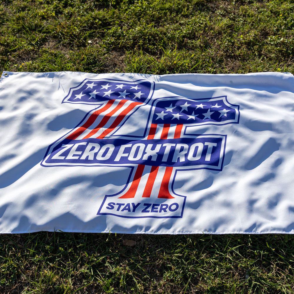 ZF Stars and Stripes Flag displayed on grass, featuring a bold blue, red, and white Zero Foxtrot emblem with stars, stripes, and Stay Zero text