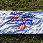 ZF Stars and Stripes Flag displayed on grass, featuring a bold blue, red, and white Zero Foxtrot emblem with stars, stripes, and Stay Zero text