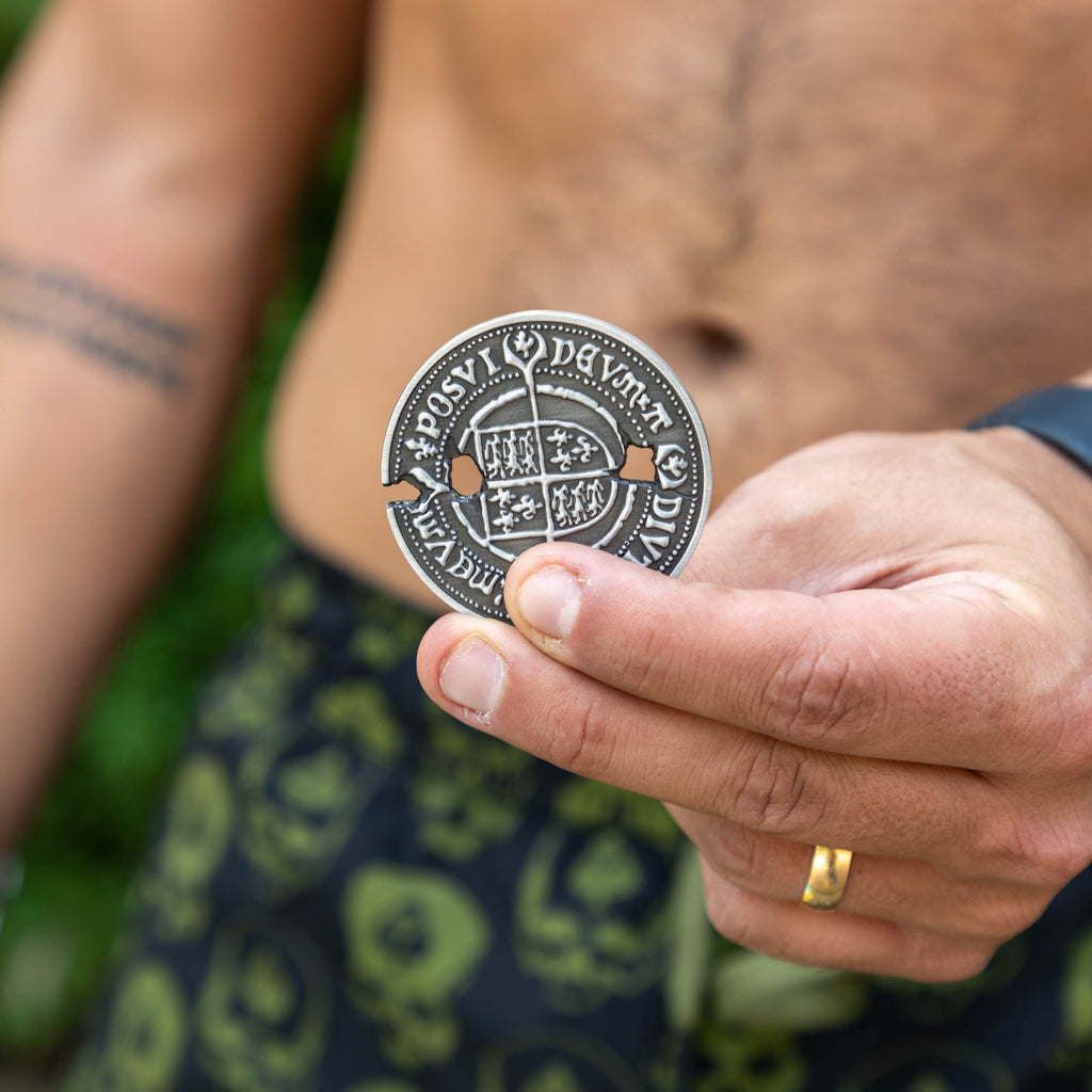 Never Say Die Coin held between fingers showing an ornate silver design with raised lettering and detailed central artwork outdoors
