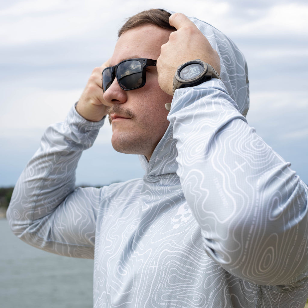 Hooded Rash Guard (Last Chance) in a light gray contour line print, worn with the hood up by a man adjusting sunglasses outdoors near the water