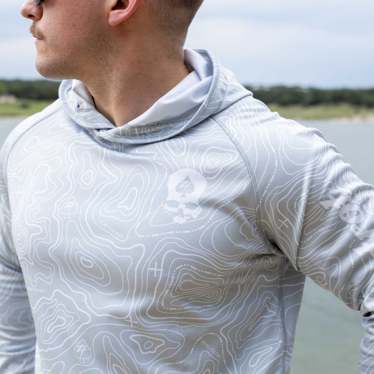 Hooded Rash Guard (Last Chance) in light gray with a white contour line pattern, shown on a person wearing the hooded long sleeve design outdoors by the water