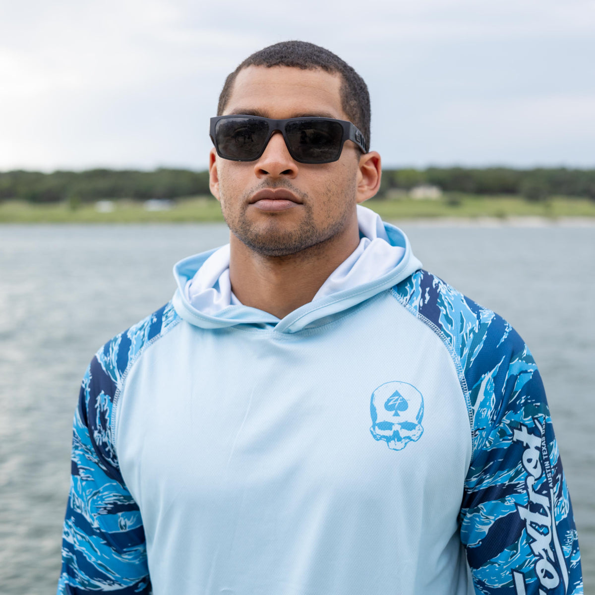 Hooded Rash Guard (Last Chance) in light blue with blue patterned sleeves, modeled outdoors by the water while wearing sunglasses