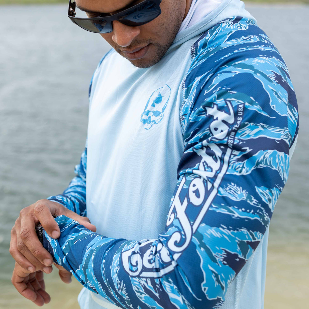 Hooded Rash Guard (Last Chance) with blue patterned sleeves and white torso, worn by a man adjusting the sleeve outdoors near the water