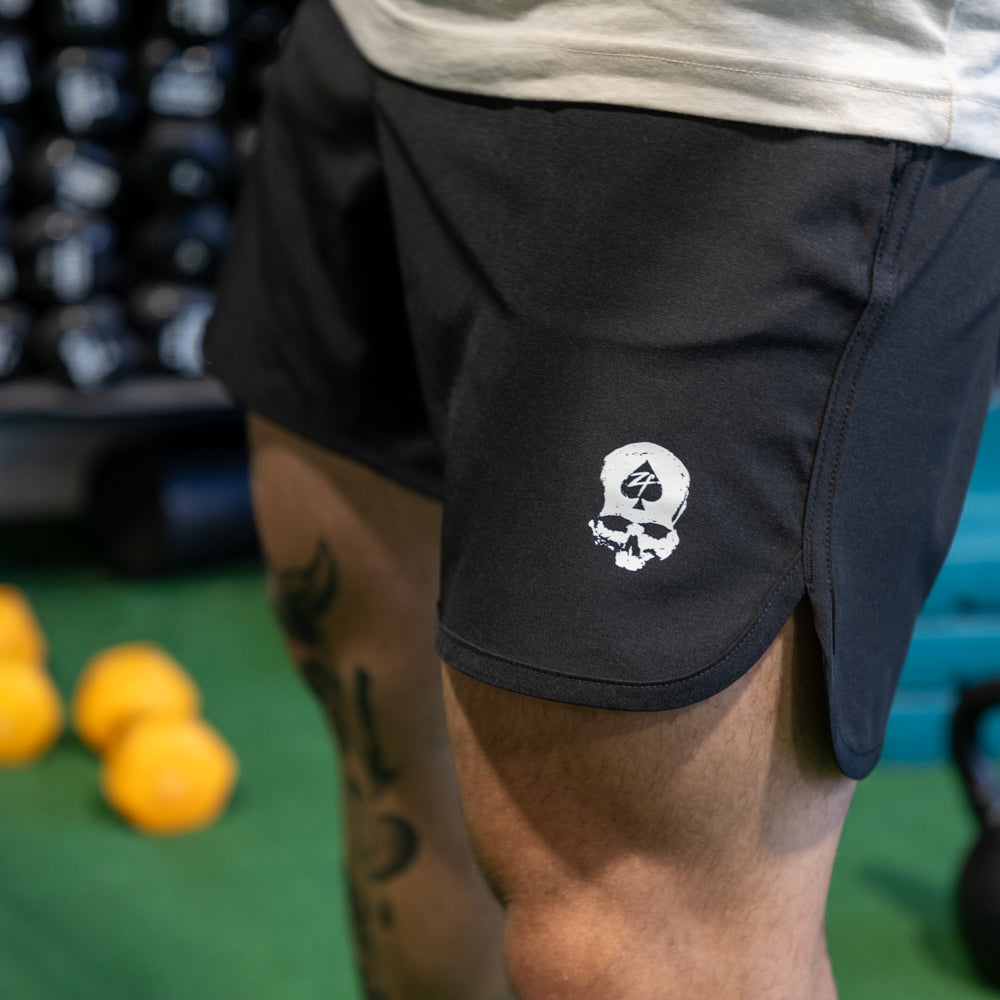 Close-up view of a person wearing black Zero Shorts - 7" Inseam with a white skull and spade logo on the left leg, standing in a gym setting with workout equipment in the background.
