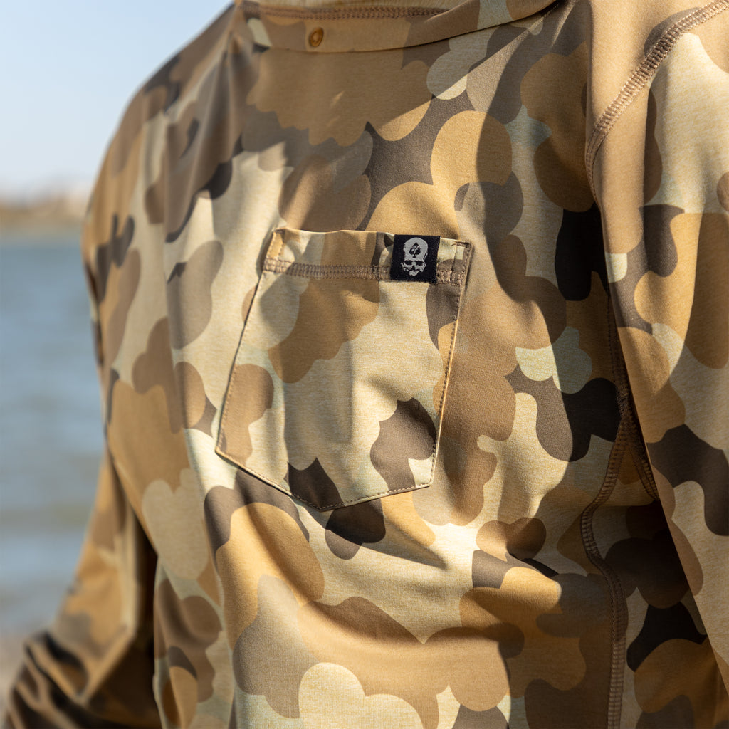 Close-up of a camo patterned Performance Sun Hoodie featuring a small chest pocket with a skull logo tag, shown outdoors near water.