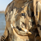 Close-up of a camo patterned Performance Sun Hoodie featuring a small chest pocket with a skull logo tag, shown outdoors near water.