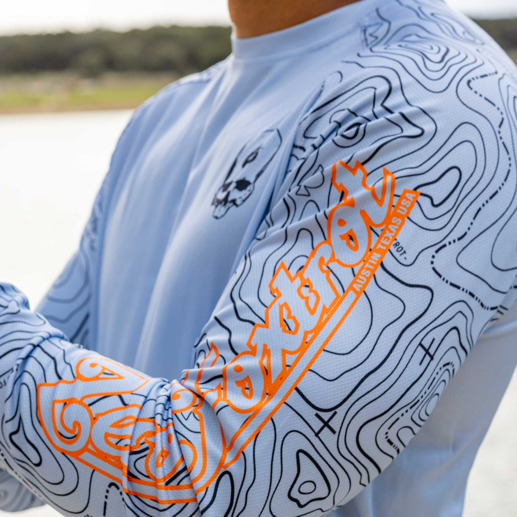 Close-up of a person wearing a light blue Crew Rash Guard (Last Season) with black topographic line patterns and bright orange text on the sleeve, featuring a small skull design on the chest.