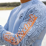 Close-up of a person wearing a light blue Crew Rash Guard (Last Season) with black topographic line patterns and bright orange text on the sleeve, featuring a small skull design on the chest.