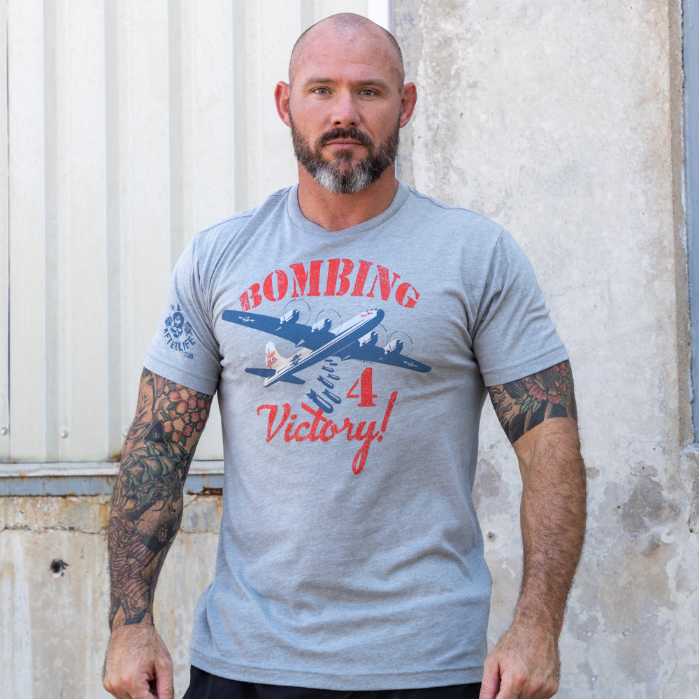 Bombing For Victory Tee in gray with vintage airplane artwork, red lettering, and a classic military inspired graphic modeled by a man outdoors