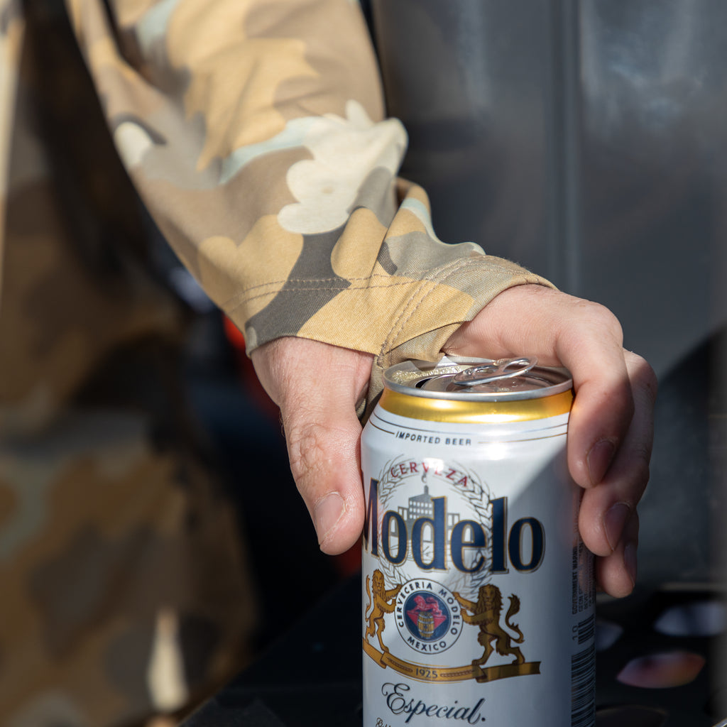Close-up of a person wearing a camo-patterned Performance Sun Hoodie holding a can of Modelo Especial beer outdoors on a sunny day.