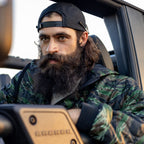 Woobie Zip Hoodie in camouflage worn by a bearded man seated in a vehicle, showing the hood, full zip front, and rugged outerwear design