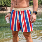 Close-up of a man standing near water wearing Swim Trunks with vertical red, white, and blue stripes and a beige drawstring waistband.