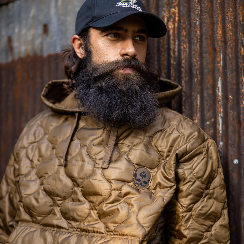 Woobie Hoodie (Last Chance) worn by a bearded man, featuring a brown quilted design, hood, drawstrings, and skull patch against a weathered metal wall