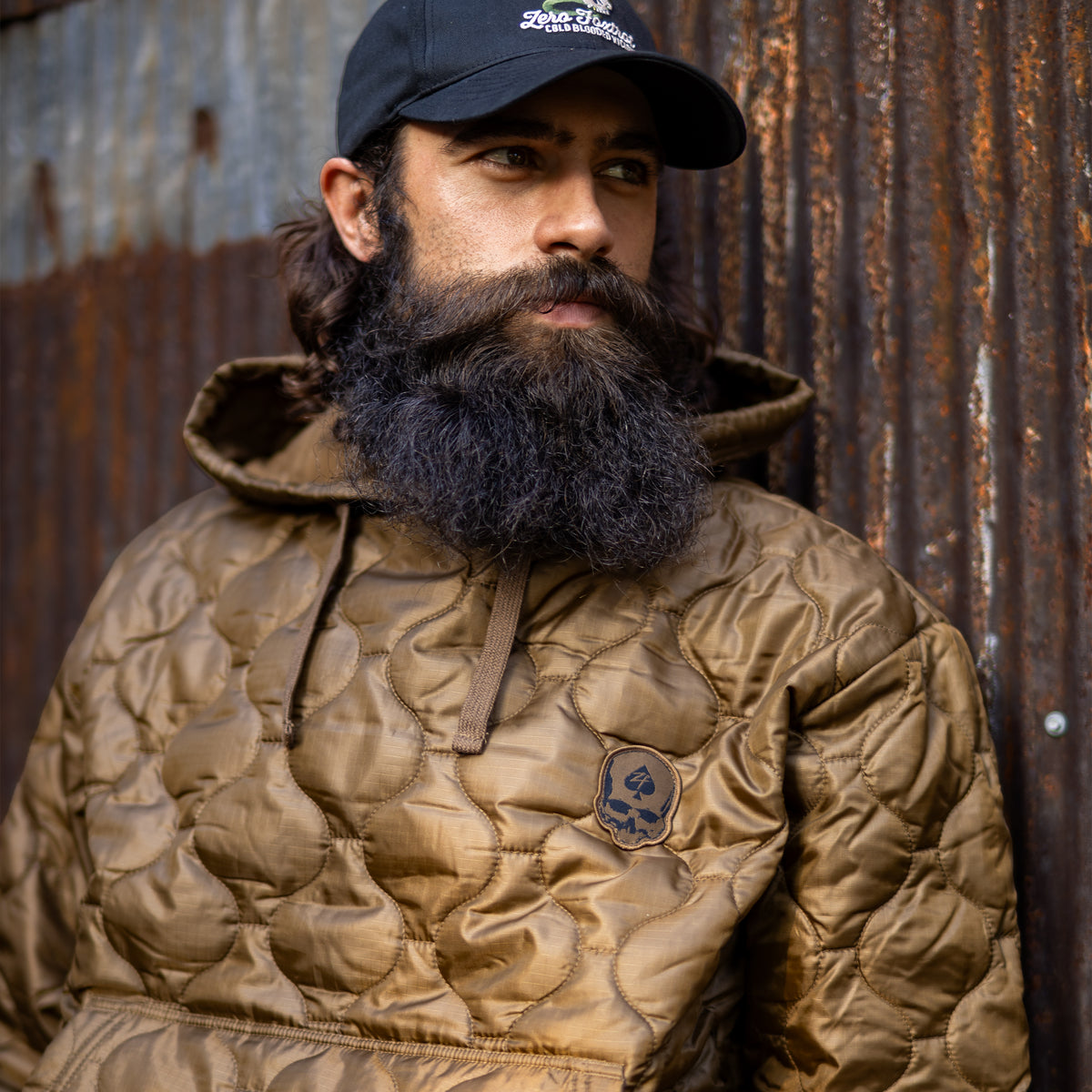 Woobie Hoodie (Last Chance) worn by a bearded man, featuring a brown quilted design, hood, drawstrings, and skull patch against a weathered metal wall