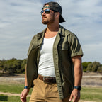 Man wearing a white Skivvy Tank under an open olive green short-sleeve shirt, sunglasses, and a backwards baseball cap standing outdoors in a natural setting.