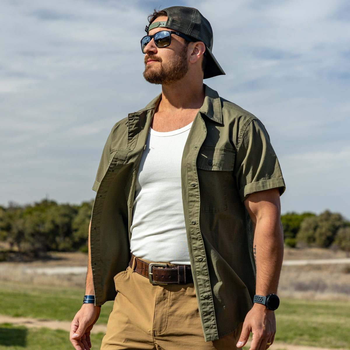 Man wearing a white Skivvy Tank under an open olive green short-sleeve shirt, sunglasses, and a backwards baseball cap standing outdoors in a natural setting.
