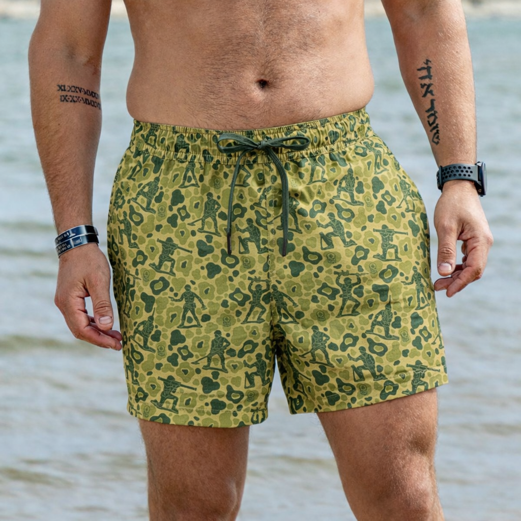 Close-up of a man wearing Army Men Swim Trunks with a green camouflage pattern featuring small soldier silhouettes, standing by the shore with water in the background.