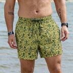 Close-up of a man wearing Army Men Swim Trunks with a green camouflage pattern featuring small soldier silhouettes, standing by the shore with water in the background.