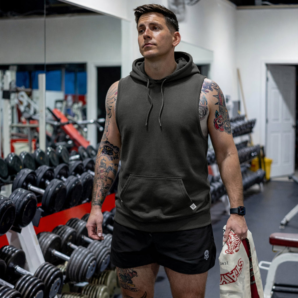 Man wearing a dark sleeveless Smuggler French Terry Muscle Hoodie standing in a gym with tattooed arms, holding a towel, surrounded by weightlifting equipment.