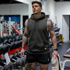 Man wearing a dark sleeveless Smuggler French Terry Muscle Hoodie standing in a gym with tattooed arms, holding a towel, surrounded by weightlifting equipment.