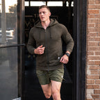 Man wearing an olive green Smuggler French Terry Zip Hoodie jogging outdoors near a brick wall and glass door on a sunny day.