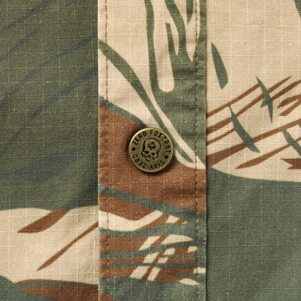 Close-up of a patterned fabric with green, brown, and beige tones featuring a metal button on the Chore Jacket with subtle texture and stitching detail.