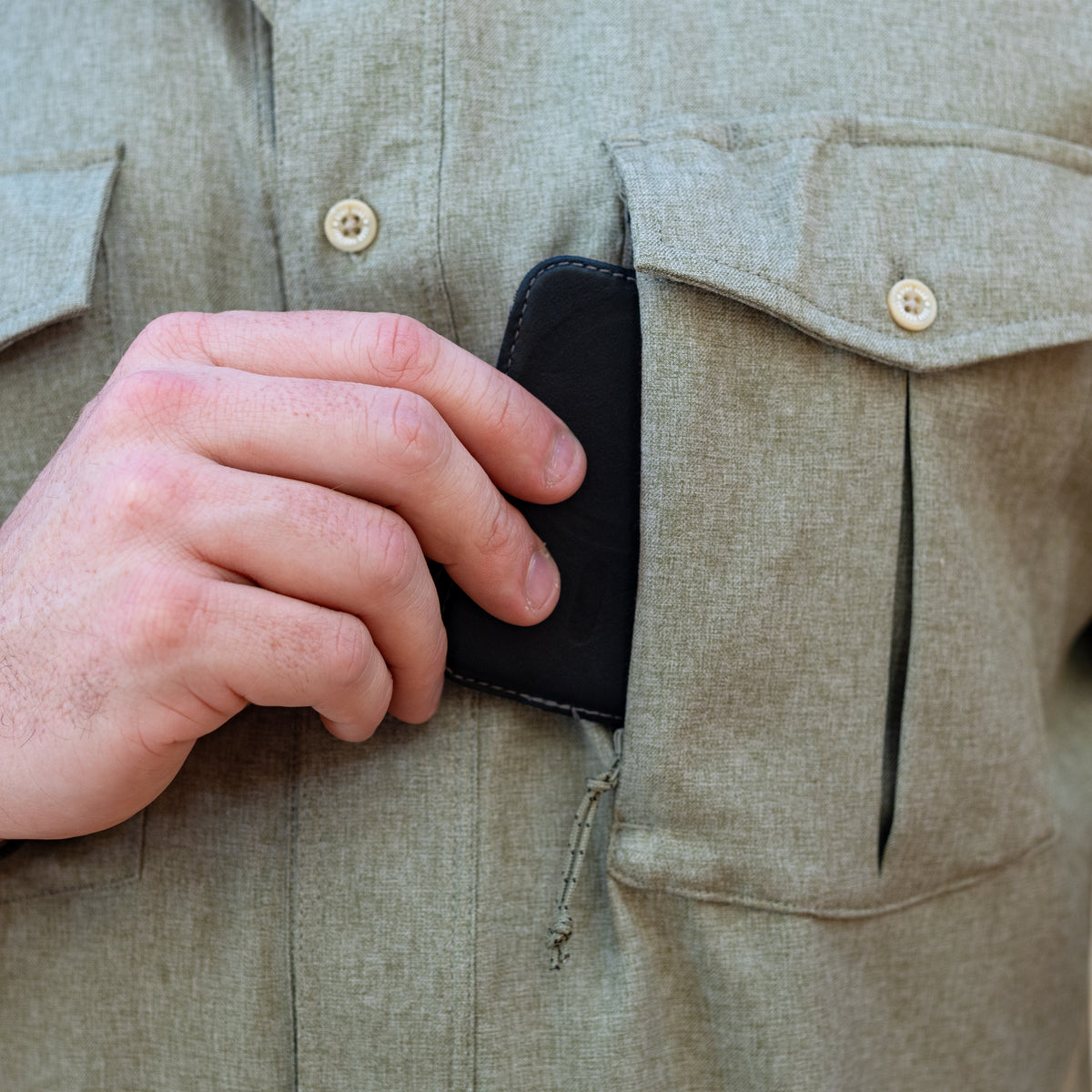 Close-up of a hand placing a black wallet into the chest zipper pocket of a light olive green F'n Soft Daily Shirt, highlighting the shirt's functional design and button details.