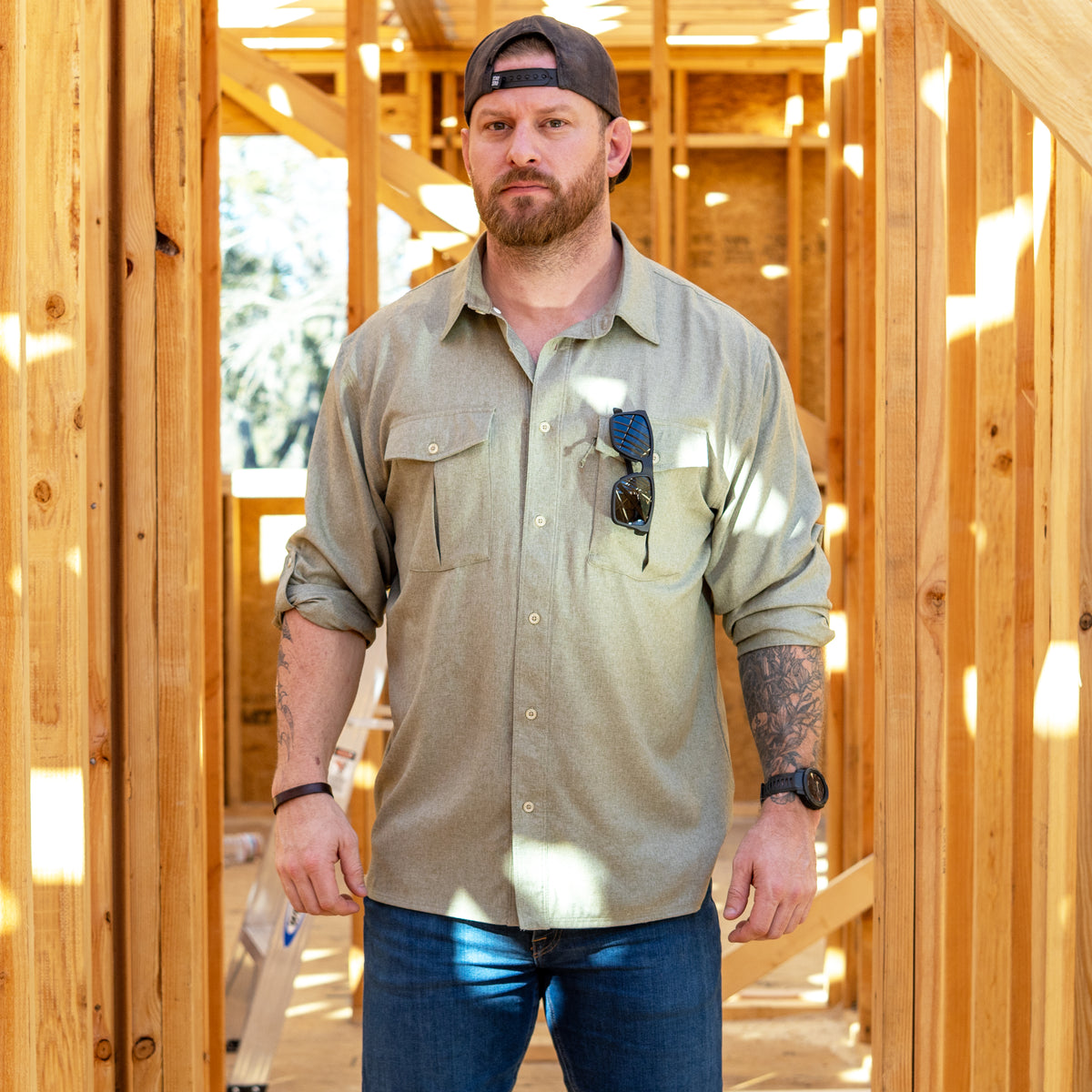 Man wearing a light-green F'n Soft Daily Shirt with rolled-up sleeves and sunglasses tucked into the chest pocket, standing inside a wooden frame structure under construction.