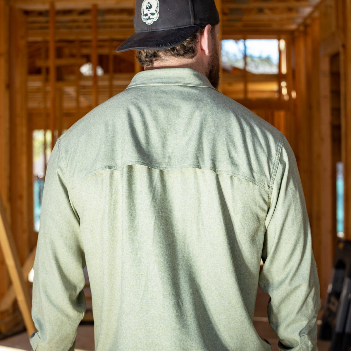 Man wearing a light green F'n Soft Daily Shirt seen from the back inside a wooden frame construction site, paired with a black cap featuring a white skull design.