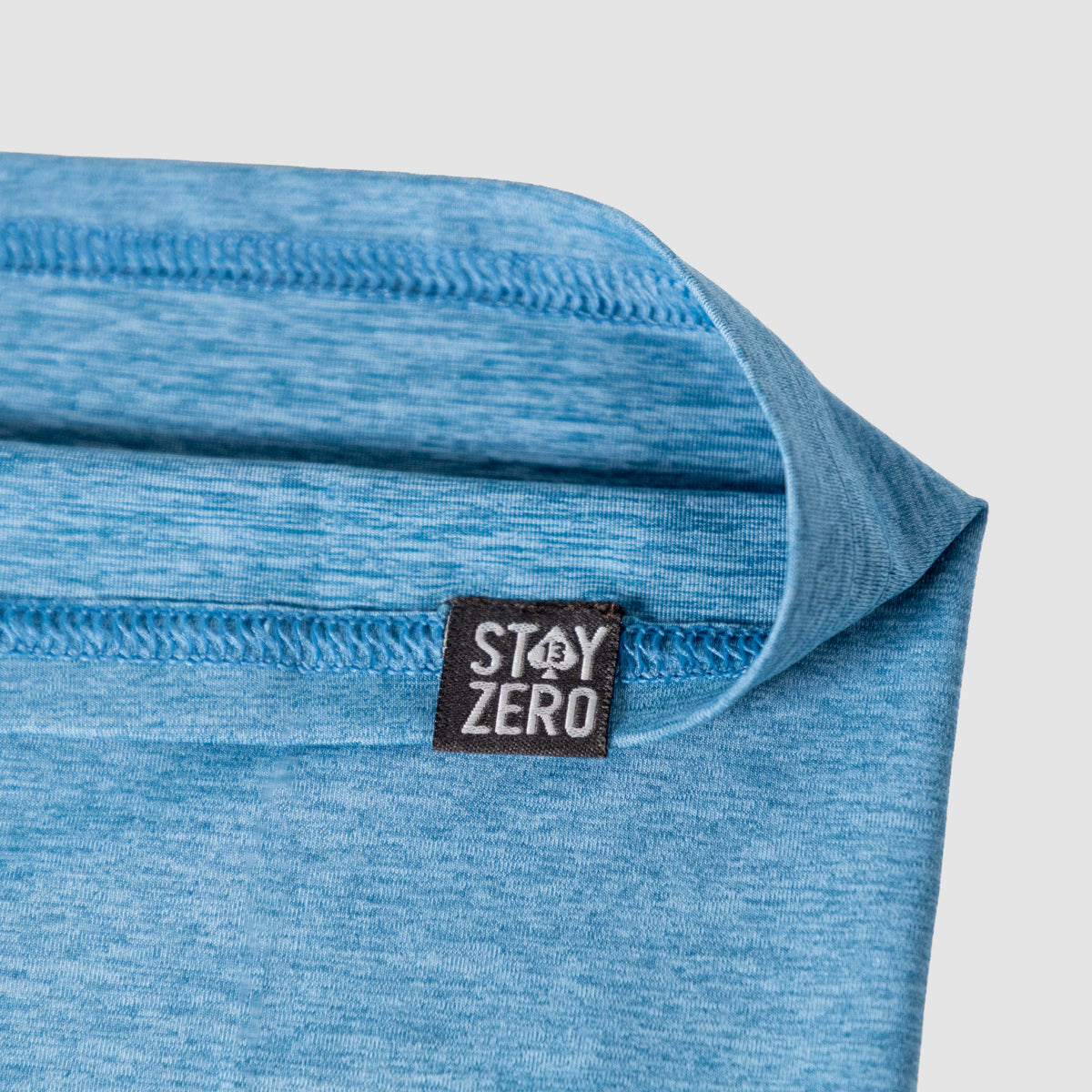 Close-up of light blue fabric with visible stitching and a black and white Stay Zero logo tag on a Performance Sun Gaiter designed for sun protection and comfort.