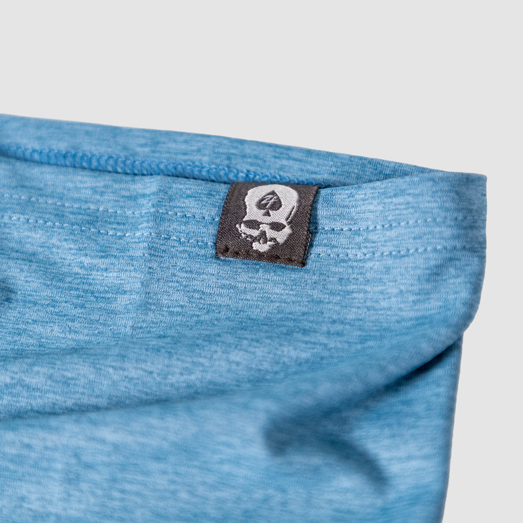 Close-up of a light blue fabric edge with a black tag featuring a white skull and spade logo, part of the Performance Sun Gaiter accessory.