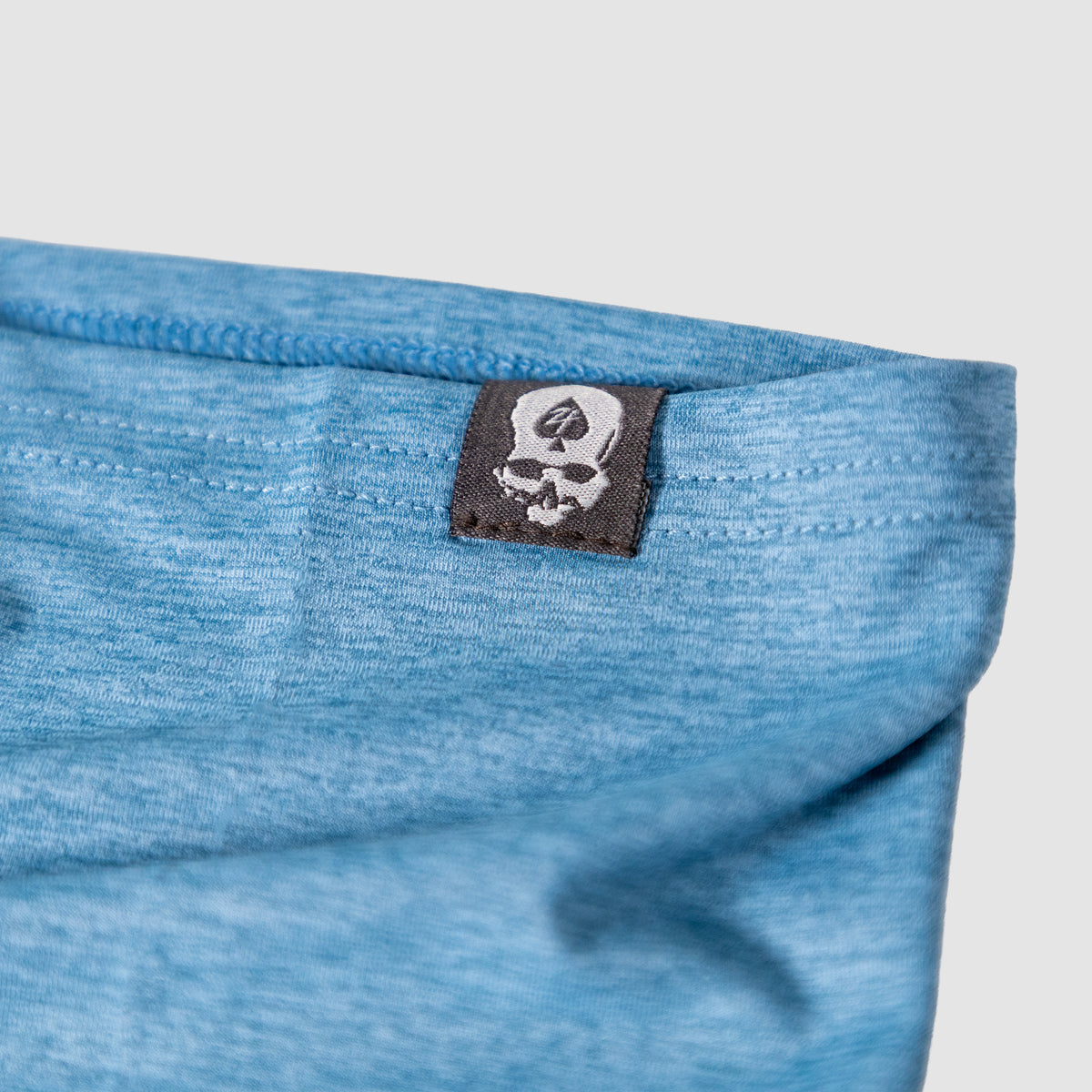 Close-up of a light blue fabric edge with a black tag featuring a white skull and spade logo, part of the Performance Sun Gaiter accessory.