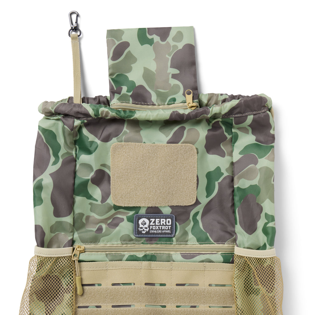 TAC Drawstring Pack in green and brown camouflage pattern with beige accents, featuring mesh side pockets, a zipper pocket, a Velcro patch area, and a metal clip attachment.
