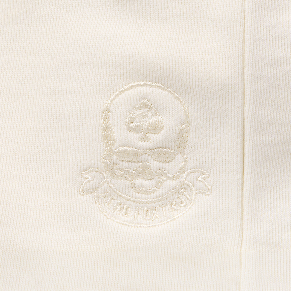 Close-up of an embroidered crest detail in matching bone white thread on the fabric of the Smuggler French Terry Zip Hoodie, highlighting its subtle texture and premium stitching.