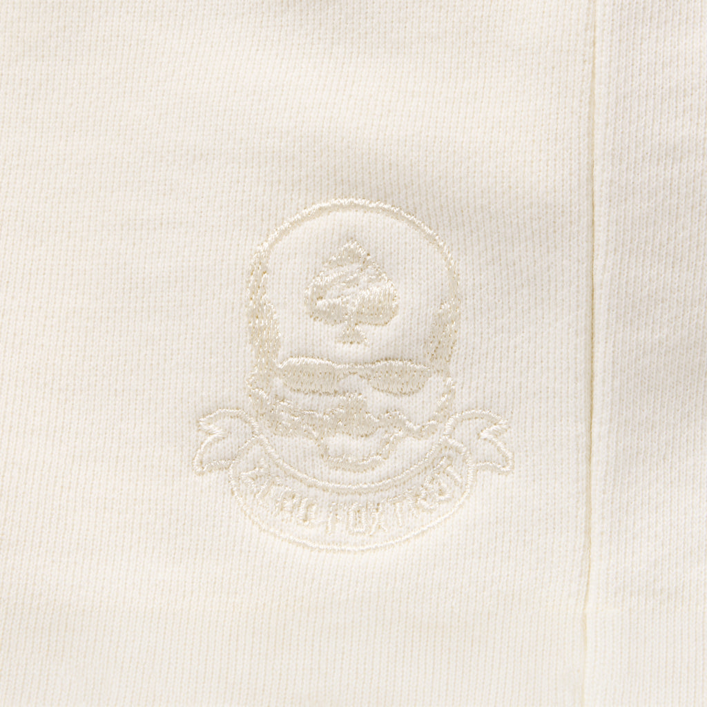 Close-up of the embroidered crest detail in matching bone white thread on the Smuggler French Terry Muscle Hoodie, showcasing subtle textured stitching on soft fabric.
