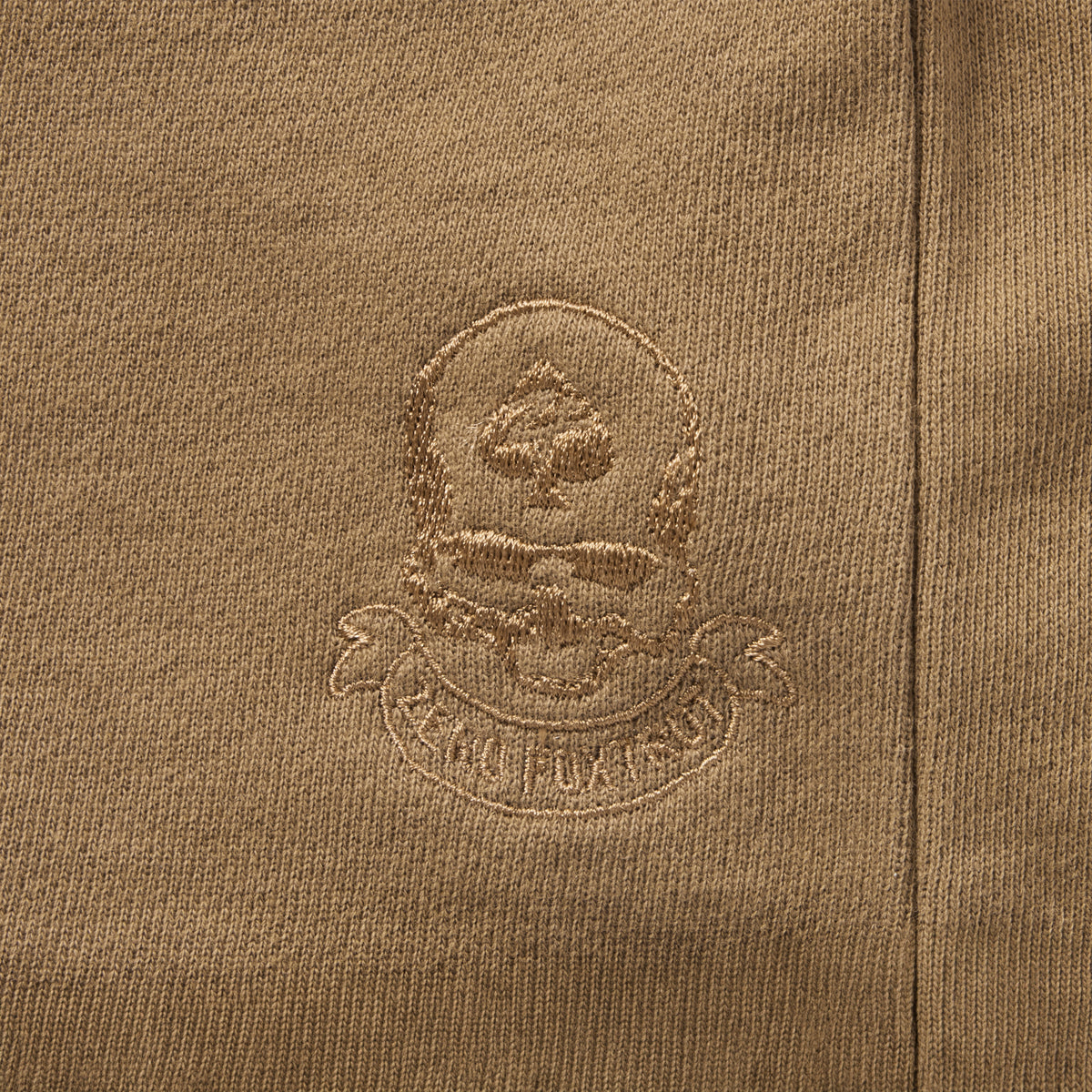 Close-up of embroidered skull and spade emblem on brown fabric of Smuggler French Terry Zip Hoodie, highlighting detailed stitching and textured material.