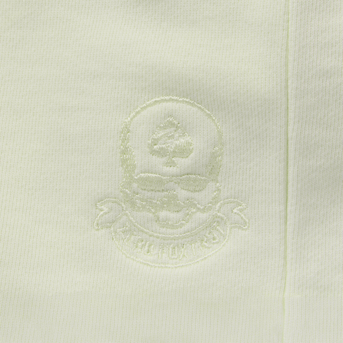 Close-up of a light sage green Smuggler French Terry Hoodie featuring a subtle embroidered skull and ace of spades logo on soft French terry fabric.